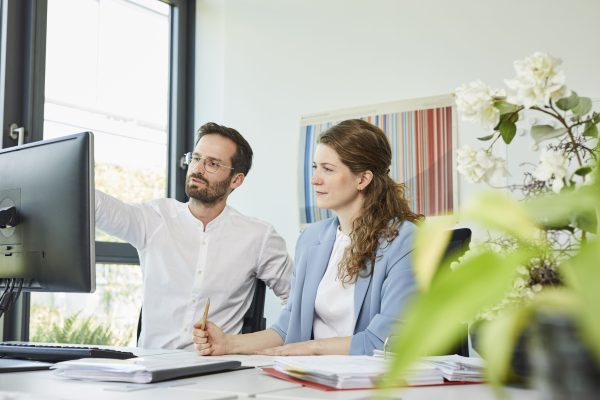 Auf dem Bild sind eine Frau und ein Mann zu sehen. Sie schauen seitlich auf einen Gegenstand, der nicht mehr im Bild zu erkennen ist. Der Mann trägt ein weißes Hemd und eine Brille und zeigt mit ausgestrecktem Arm auf etwas außerhalb des Bildrandes. Die Frau hat lange, braune Haare und trägt einen hellblauen Blazer. Darunter trägt sie ein weißes Oberteil. Auf dem Tisch vor ihnen liegen verschiedene Arbeitsmaterialien.